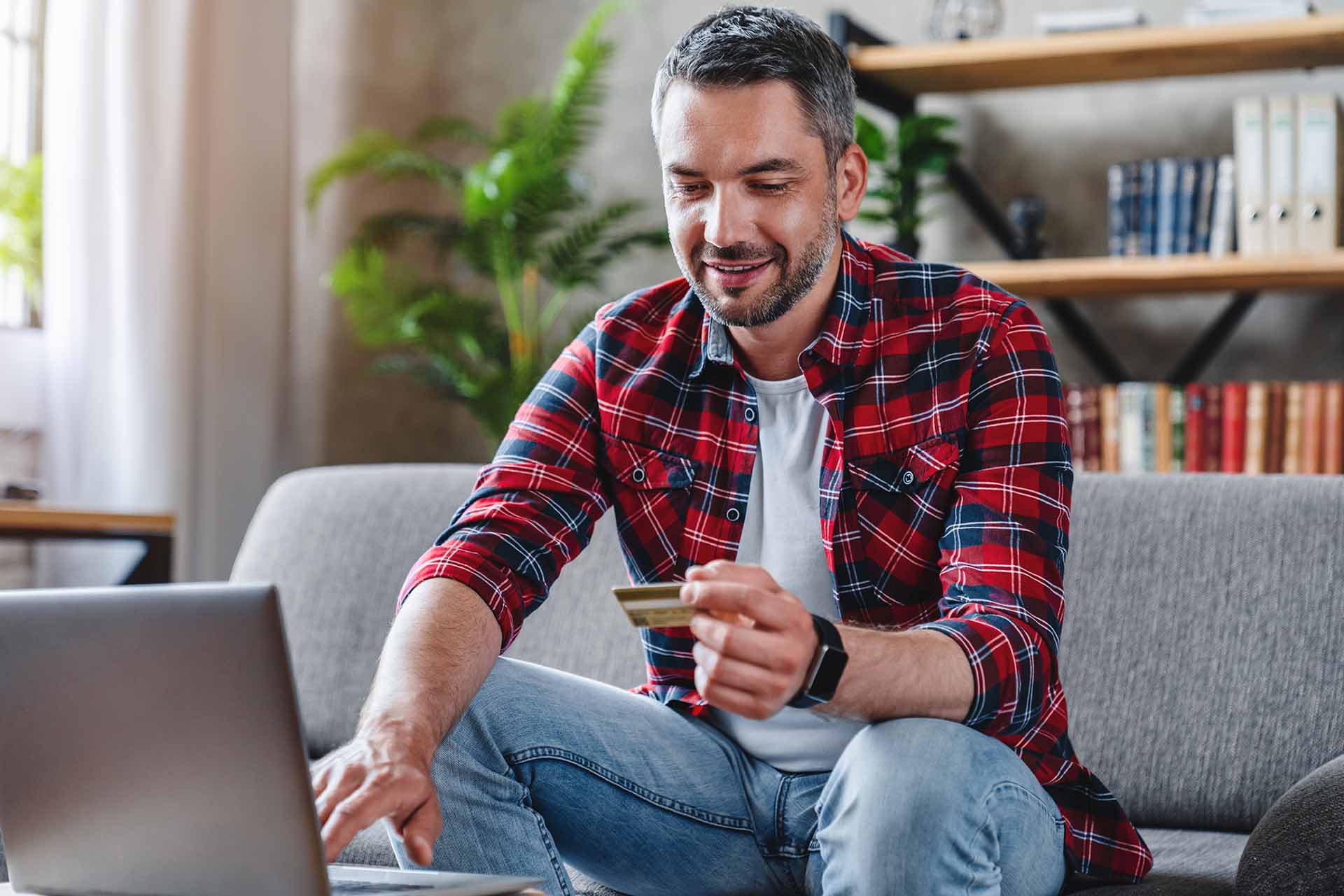 Hispanic male sitting on couch in home smiling and holding credit card while using laptop