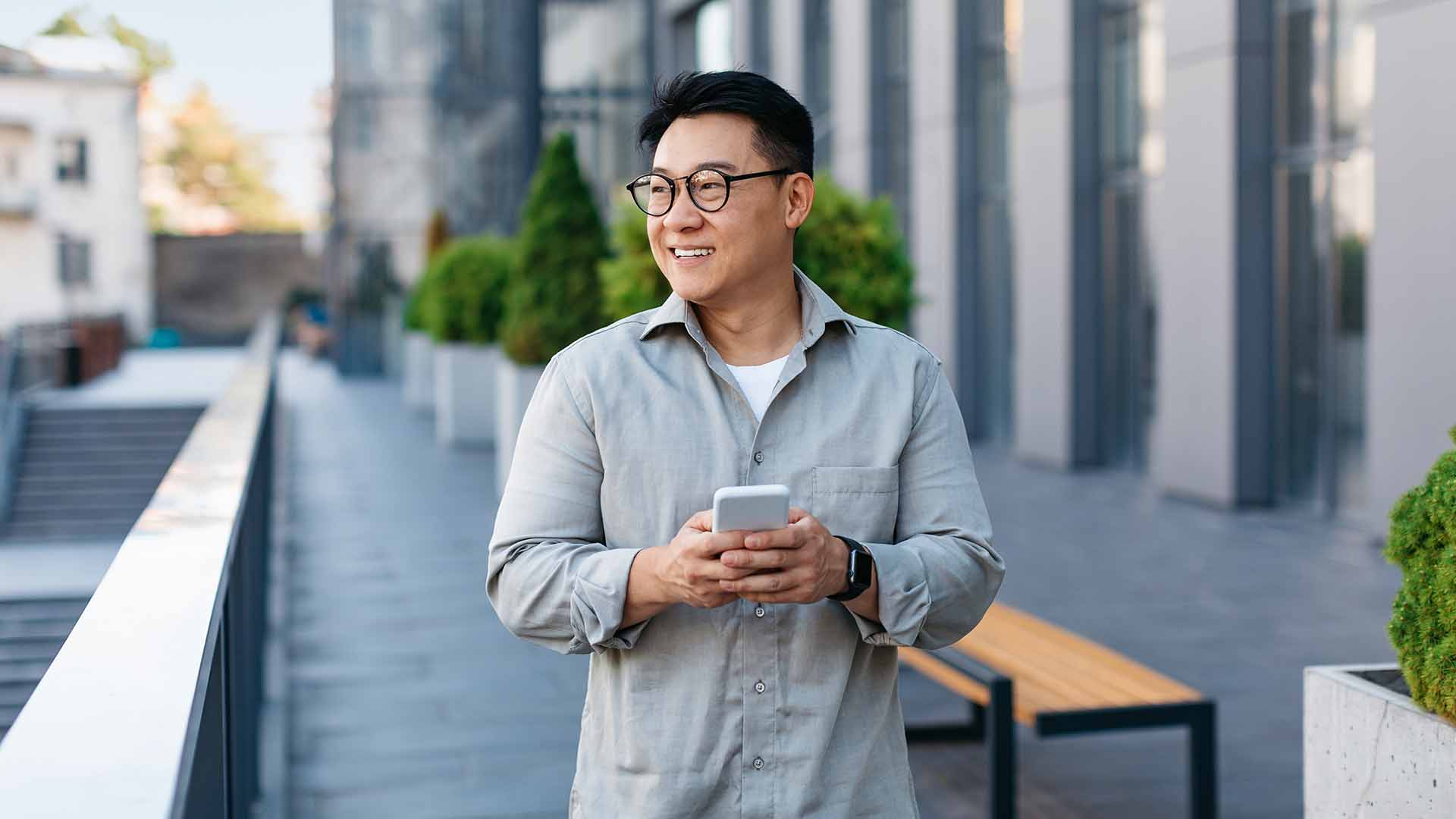 Asian man with glasses smiling while holding phone outside